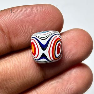 May include: A square bead with a distinctive pattern of concentric circles in red, yellow, blue, and black. The bead is held between two fingers, highlighting its intricate design and vibrant colours. The bead's dimensions are approximately 1 cm.