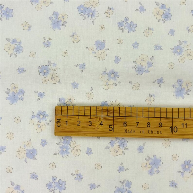 May include: White fabric featuring a floral design in pale blue and cream hues. A wooden ruler with centimetre markings is placed on the fabric, showing measurements up to 11 cm.