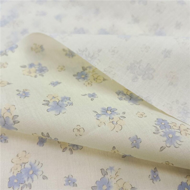 May include: Close-up of a cream fabric with a delicate floral pattern. The design features small blue and yellow flowers with grey stems and leaves scattered across the surface. The fabric is folded, revealing the pattern on both sides.