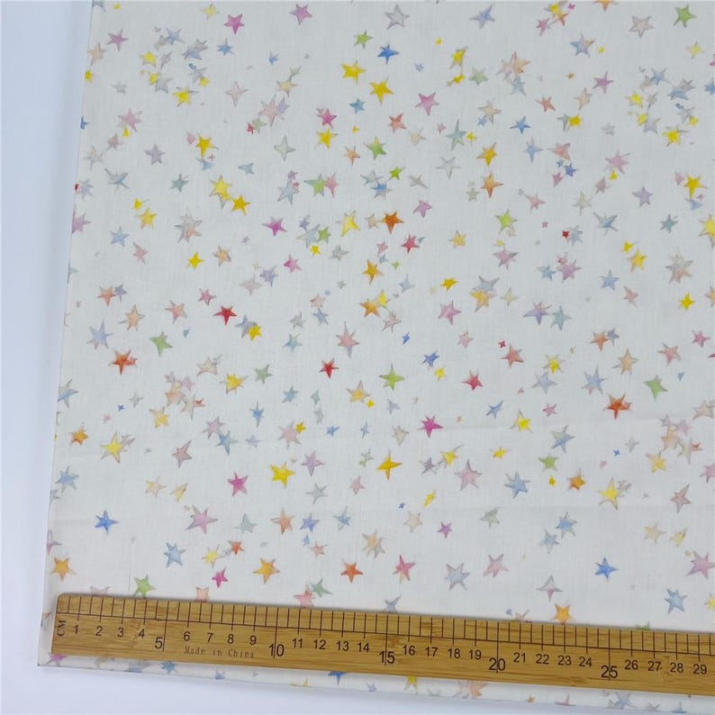 May include: White fabric patterned with small, multi-coloured stars. The stars are in shades of red, yellow, blue, green, and pink. A wooden ruler with centimetre markings is at the bottom.