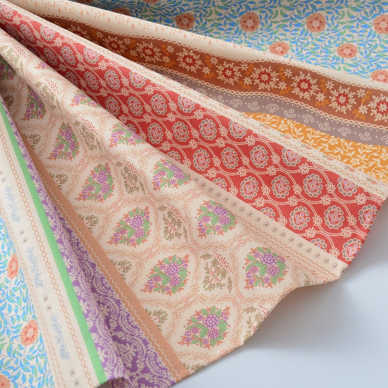 Op de afbeelding: Een veelkleurige stof met een herhalend bloemenpatroon. De stof is gevouwen op een manier die de verschillende kleuren en patronen laat zien. De stof is licht beige van kleur met strepen in rood, oranje, groen en paars.