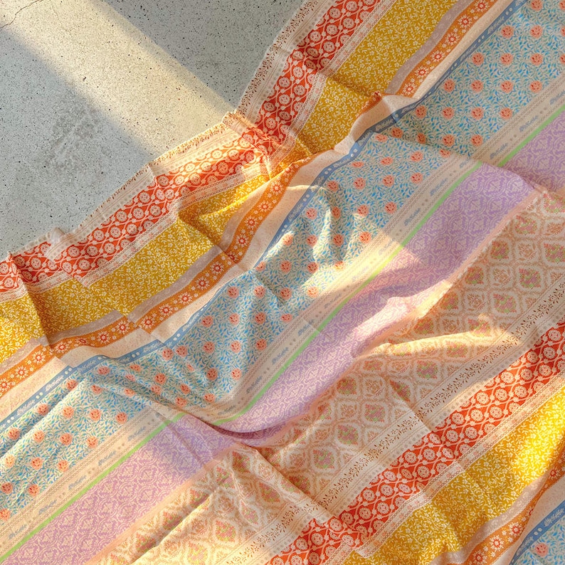 Op de afbeelding: Een kleurrijke stof met een gestreept patroon met verschillende bloemen- en geometrische ontwerpen in tinten geel, oranje, roze, blauw en wit.