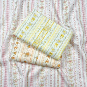 May include: Two pieces of fabric with floral patterns. The top fabric is light blue with yellow and white floral patterns and bows. The bottom fabric is white with orange and yellow floral patterns. Both fabrics have a striped background.
