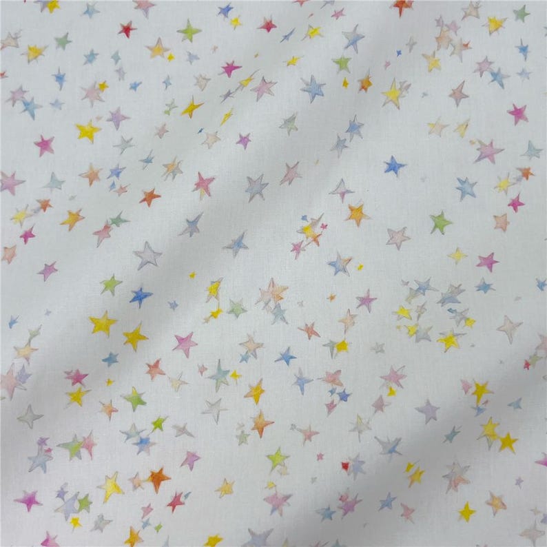May include: White fabric featuring a pattern of watercolour stars in multiple colours, such as yellow, blue, pink, and orange. The stars vary in size and are spread across the fabric. The fabric has a soft, slightly textured appearance.