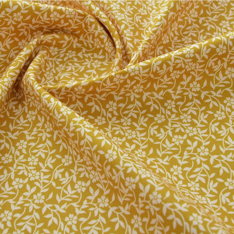 Op de afbeelding: Een gele stof met een wit bloemenpatroon. De stof heeft een kleinbloemig print met witte bloemen en bladeren op een gele achtergrond.
