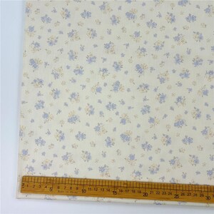 May include: Cream-coloured fabric featuring a repeating pattern of small blue and tan floral clusters. A wooden ruler with centimetre markings is placed along the bottom edge of the fabric. The ruler shows measurements up to 30 cm.