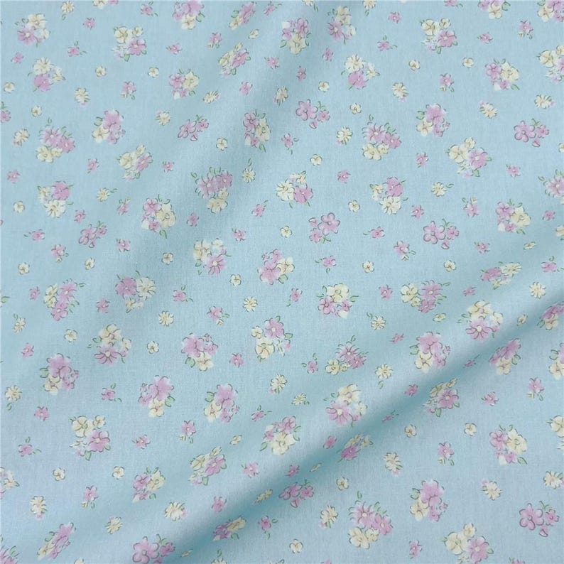 May include: Pale blue fabric featuring a delicate floral design. The pattern consists of small clusters of pink and cream-coloured flowers. The fabric appears soft and slightly creased, possibly made from cotton or a similar natural fibre.