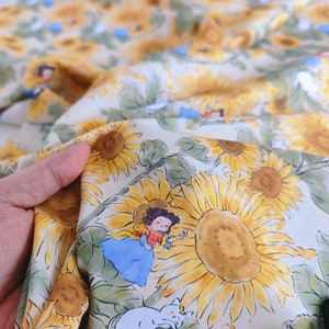 Puede incluir: Una tela beige con un patr&oacute;n repetitivo de girasoles y una ni&ntilde;a con un gato. La ni&ntilde;a lleva un vestido azul y un sombrero de paja. Los girasoles son amarillos con centros marrones.