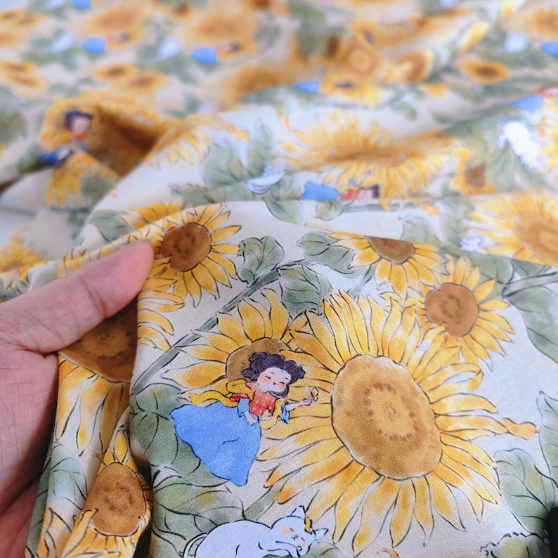 Puede incluir: Una tela beige con un patr&oacute;n repetitivo de girasoles y una ni&ntilde;a con un gato. La ni&ntilde;a lleva un vestido azul y un sombrero de paja. Los girasoles son amarillos con centros marrones.