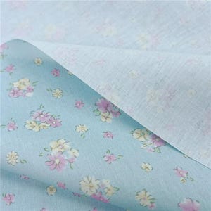 May include: Pale blue floral fabric featuring a pattern of pink and yellow flowers. The fabric is folded, showing a plain white reverse side. The floral design is scattered across the blue surface.