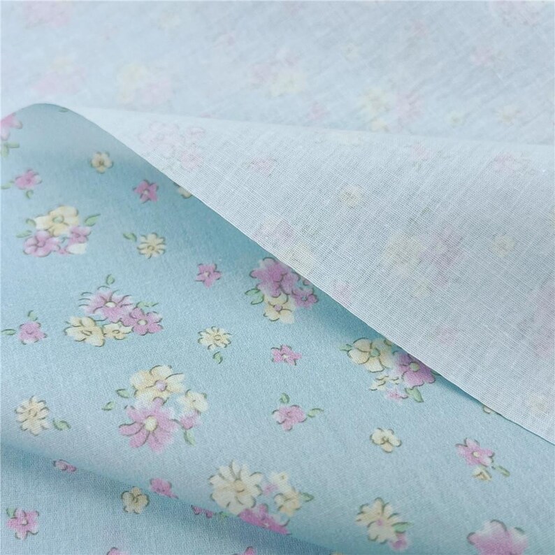 May include: Pale blue floral fabric featuring a pattern of pink and yellow flowers. The fabric is folded, showing a plain white reverse side. The floral design is scattered across the blue surface.