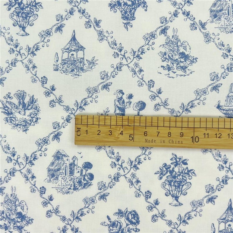 May include: White fabric with a blue toile pattern featuring floral vines, baskets, birds, and pastoral scenes. A wooden ruler shows measurements up to 13 cm. The fabric has a classic, vintage aesthetic.