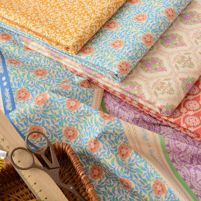 Op de afbeelding: Een stapel stof met bloemmotieven in verschillende kleuren, waaronder oranje, blauw, roze en groen. De stof is gevouwen en gerangschikt op een geweven mand met een liniaal en een schaar.