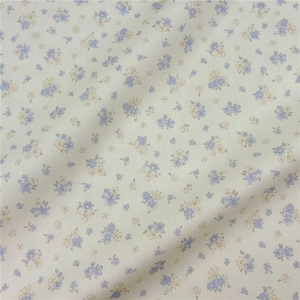 May include: A close-up shot of a fabric featuring a repeating floral design. The fabric has a cream-coloured backdrop, adorned with small clusters of blue and pale yellow flowers. The material appears soft and slightly creased.