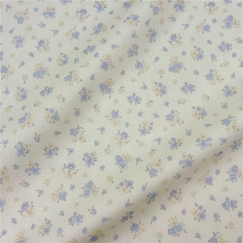 May include: A close-up shot of a fabric featuring a repeating floral design. The fabric has a cream-coloured backdrop, adorned with small clusters of blue and pale yellow flowers. The material appears soft and slightly creased.