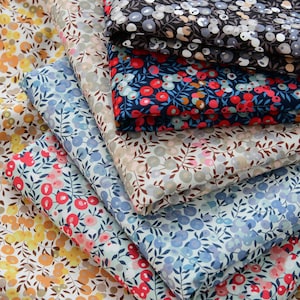 Op de afbeelding: Een stapel bloemige stofstalen in verschillende kleuren. De stoffen hebben kleine bloemen- en bladerpatronen in tinten geel, oranje, rood, blauw en grijs. De stoffen zijn gevouwen en opgestapeld, met de verschillende ontwerpen en kleuren.