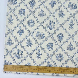 May include: Cream fabric with a blue toile pattern. The design includes scenes of gazebos, floral arrangements, fountains, and figures, linked by a trellis of vines. A ruler is placed on the fabric, showing measurements in inches.