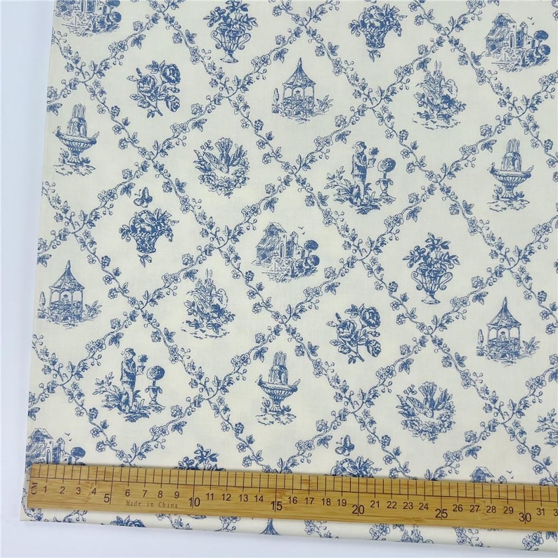 May include: Cream fabric with a blue toile pattern. The design includes scenes of gazebos, floral arrangements, fountains, and figures, linked by a trellis of vines. A ruler is placed on the fabric, showing measurements in inches.