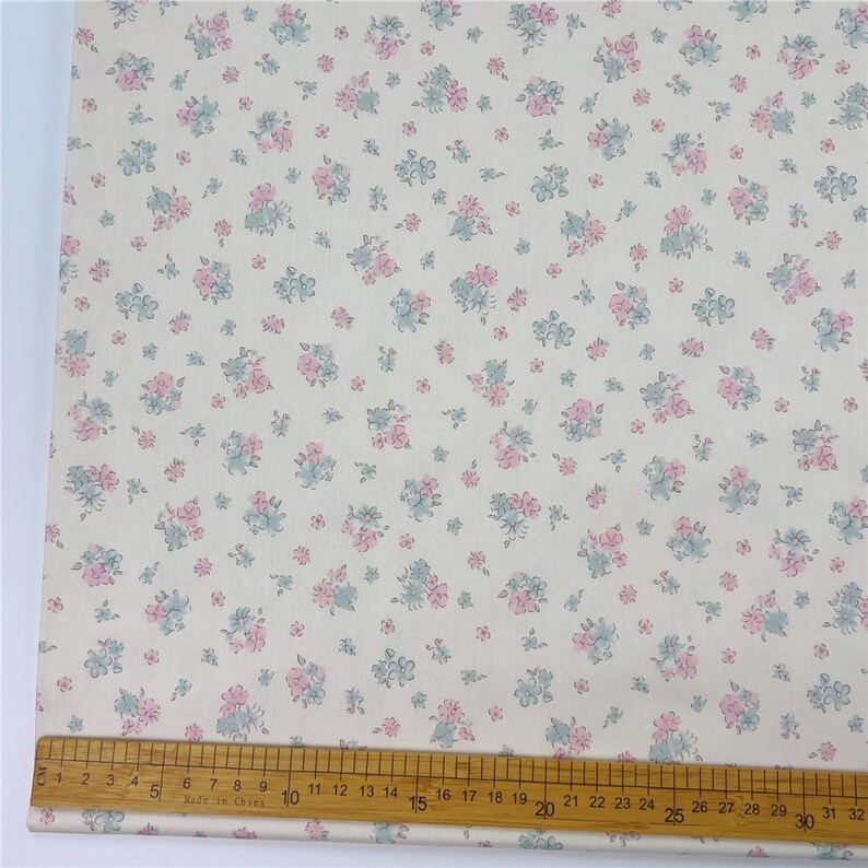 May include: Cream-coloured fabric featuring a repeating pattern of small pink and blue floral clusters. A wooden ruler marked in inches is used for measurement. The floral design is evenly distributed across the fabric's surface.