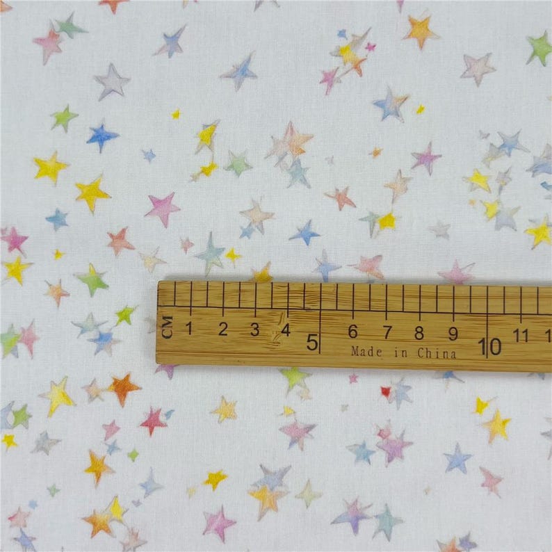 May include: White fabric patterned with multi-coloured stars in shades of yellow, pink, blue, and orange. A wooden ruler with centimetre markings is placed on the fabric. The ruler reads "Made in China".