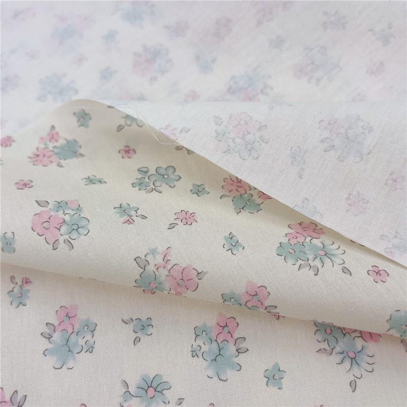 May include: Cream-coloured fabric with a floral design. The pattern includes clusters of pink and blue flowers with green stems and leaves. The fabric is folded, showing the pattern on both sides. The flowers are scattered across the material.