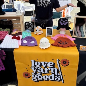 Puede incluir: Un puesto de mercado que muestra varios sombreros y figuras de fantasmas de ganchillo hechos a mano. Los sombreros son de diferentes colores y diseños, incluyendo un sombrero blanco con un lazo rojo y un sombrero negro con una cara de calabaza. La mesa tiene el texto "love yarn goods".