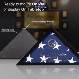 May include: A black shadow box with a folded American flag inside. The flag is displayed in a triangular shape with a silver United States Air Force emblem. The box has a sawtooth hanger installed on the back and rotating clips on the front. The text on the flag reads "Robert J. Beasley, United States Army, June 21, 1937 - May 30, 2022".