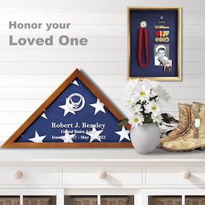 May include: A wooden shadow box with a folded American flag inside. The flag is blue with white stars and has the United States Air Force logo in the center. The box has a personalized inscription that reads "Robert J. Beasley, United States Army, June 21, 1957 - May 30, 2022".