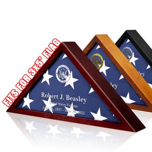 May include: Three triangular wooden flag display cases in dark cherry, light oak, and black finishes. Each case holds a folded American flag with a veteran emblem and personalized text reading Robert J. Beasley, United States Veteran, June 21, 1937 - May 30, 2022. Fits for 3x5' flag.