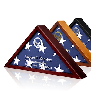 May include: Three triangular wooden flag display cases in dark cherry, light oak, and black finishes. Each case holds a folded American flag with a United States Air Force emblem and personalized text reading Robert J. Beasley, United States Army.