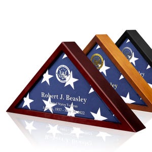 May include: Three triangular wooden flag display cases in dark cherry, light oak, and black finishes. Each case holds a folded American flag with a United States Veteran emblem and personalized text: Robert J. Beasley, United States Veteran, June 21, 1937 - May 30, 2022.
