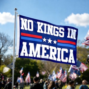 Puede incluir: Una bandera azul con las palabras blancas "NO KINGS IN AMERICA". La bandera tiene rayas rojas y azules y tres estrellas blancas. Está en un mástil plateado, con otras banderas y personas al fondo.