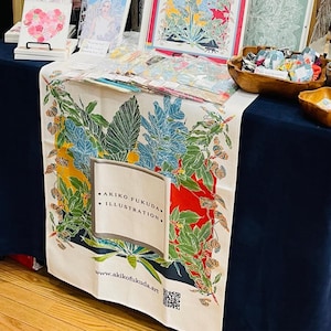May include: A stall showcasing art prints and textiles with botanical designs. A white tablecloth with a vibrant floral pattern is draped over the table. The text "AKIKO FUKUDA ILLUSTRATION" is printed on the cloth. Framed art and wooden bowls are also visible.