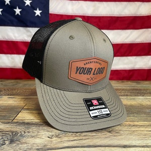 Puede incluir: Una gorra de camionero verde y negra con un parche de cuero marr&oacute;n que dice "Brentsbrig Your Logo 20 x 21". La gorra est&aacute; sobre una superficie de madera delante de una bandera estadounidense.