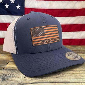 May include: A navy blue trucker hat with a white mesh back. The hat features a brown leather patch with an American flag design and the text "CUSTOM TEXT".