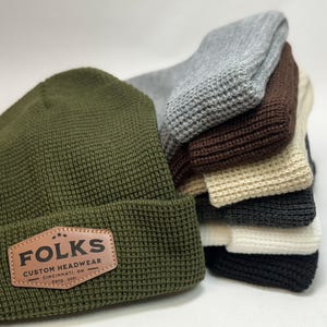 May include: An olive green beanie with a brown leather patch that reads "FOLKS CUSTOM HEADWEAR CINCINNATI, OH ESTD. 1991". A stack of other beanies in gray, brown, beige, dark gray, and white are behind it. The beanies have a waffle knit texture.
