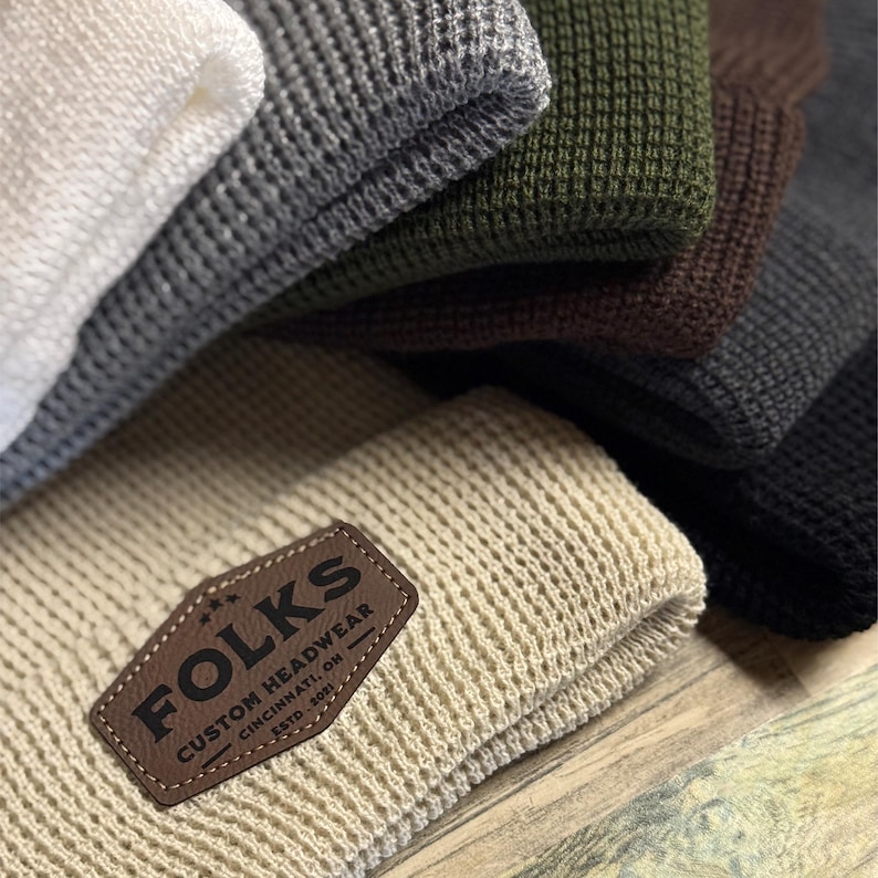 May include: Close-up of a stack of knit beanies in various colors, including white, gray, olive green, brown, and black. The front beanie is beige with a brown leather patch that reads "FOLKS CUSTOM HEADWEAR CINCINNATI, OH ESTD. 2021."