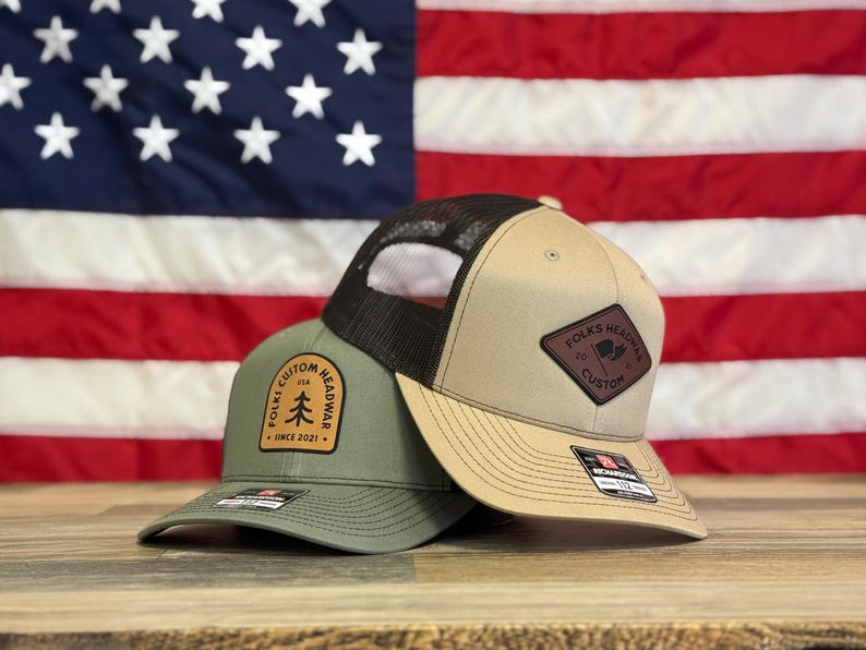 Puede incluir: Dos gorras de camionero, una verde y otra beige, con una malla trasera marr&oacute;n. La gorra verde tiene un parche de cuero marr&oacute;n con el texto "Folks Custom Headwear USA Since 2021" y un peque&ntilde;o gr&aacute;fico de un pino. La gorra beige tiene un parche de cuero marr&oacute;n con el texto "Folks Headwear Co. Custom" y un peque&ntilde;o gr&aacute;fico de una bandera.