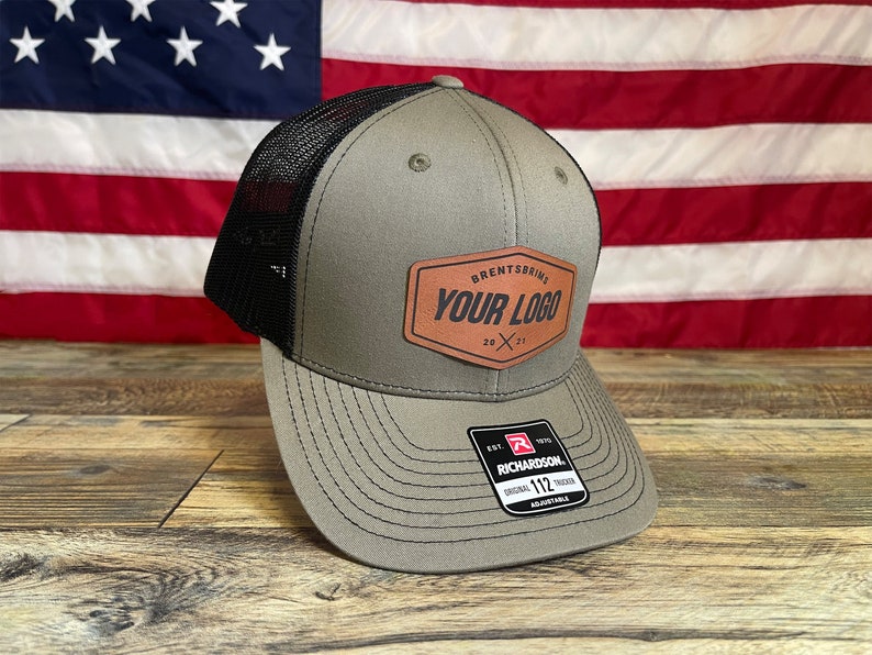 Puede incluir: Una gorra de camionero verde y negra con un parche de cuero marr&oacute;n que dice "Brentsbrig Your Logo 20 x 21". La gorra est&aacute; sobre una superficie de madera delante de una bandera estadounidense.