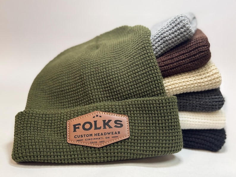 May include: An olive green beanie with a textured waffle knit design and a brown leather patch that reads "FOLKS CUSTOM HEADWEAR CINCINNATI, OH ESTD. 2021". A stack of beanies in various colors is behind it.