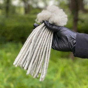 May include: A bundle of gray synthetic dreadlocks, held in a gloved hand. The dreadlocks are long and straight, with a slightly textured surface.