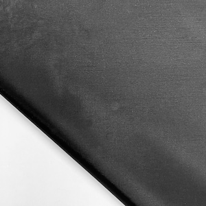 May include: A close-up shot of a black fabric with a subtle texture. The fabric is folded diagonally across the frame, with a white background visible in the lower left corner. The material appears smooth and possibly silky.