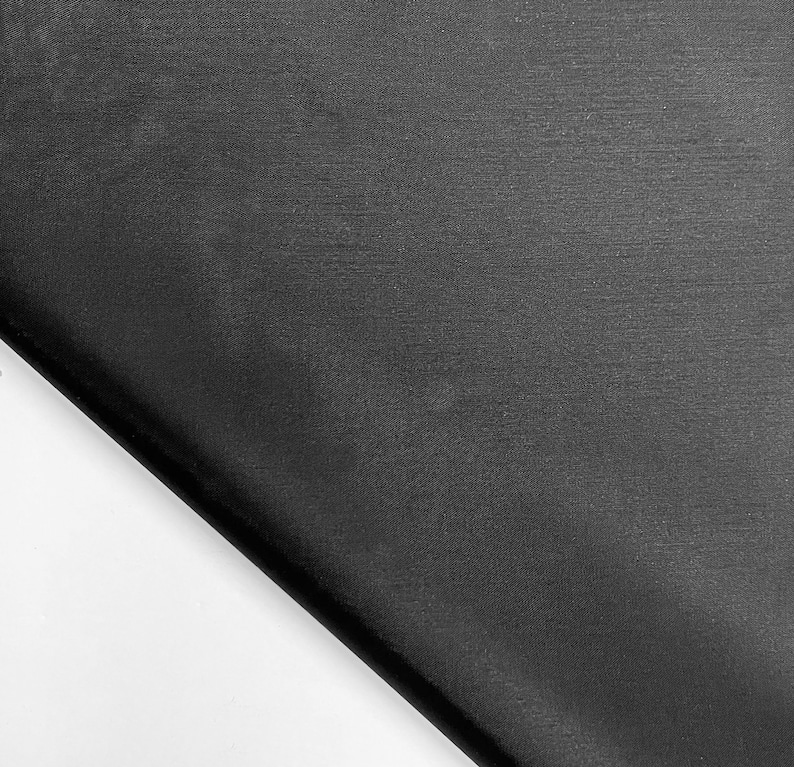 May include: A close-up shot of a black fabric with a subtle texture. The fabric is folded diagonally across the frame, with a white background visible in the lower left corner. The material appears smooth and possibly silky.