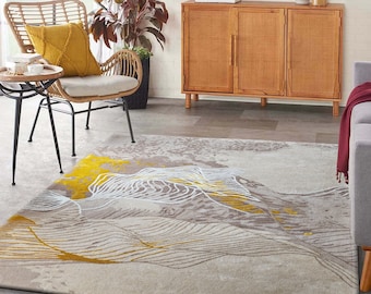 May include: A beige area rug with a mountain-like design in shades of gray, yellow, and white. The rug features a textured pattern and is placed in a living room setting with a wicker chair and wooden cabinet.