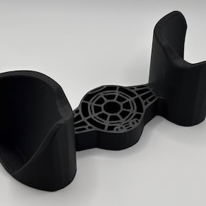 May include: A black 3D-printed device with a unique design. It features two curved holders on either side and a central, patterned section. The object is made of a matte black material and appears to be a holder or stand for an electronic device.