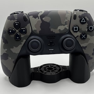 Peut inclure: Une manette de jeu vidéo à motif camouflage posée sur un support noir. La manette présente un mélange de couleurs grises, noires et vert olive. Le support a un design circulaire au centre et deux pieds de support.