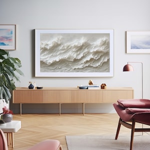 May include: A modern living room interior with a large framed artwork depicting abstract white waves hanging on a light gray wall above a light wood cabinet. The cabinet has three drawers and is sitting on four gold metal legs. A pink armchair with a red cushion is in the foreground.