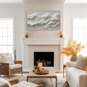 May include: A white brick fireplace with a black fireplace insert. A white couch with a throw pillow sits in front of the fireplace. A wooden coffee table with a tray of candles and decorative items sits in front of the couch. A white wicker armchair sits to the left of the coffee table. A large framed abstract painting of white waves hangs above the fireplace.