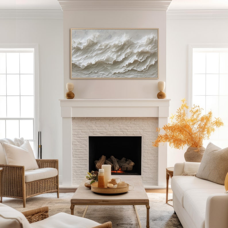 May include: A white brick fireplace with a black fireplace insert. A white couch with a throw pillow sits in front of the fireplace. A wooden coffee table with a tray of candles and decorative items sits in front of the couch. A white wicker armchair sits to the left of the coffee table. A large framed abstract painting of white waves hangs above the fireplace.