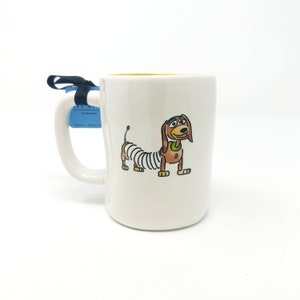 Puede incluir: Taza de cer&aacute;mica blanca con un borde amarillo que presenta una ilustraci&oacute;n de dibujos animados de Slinky Dog de la franquicia Toy Story. El perro es marr&oacute;n con un cuerpo de resorte y un collar verde.